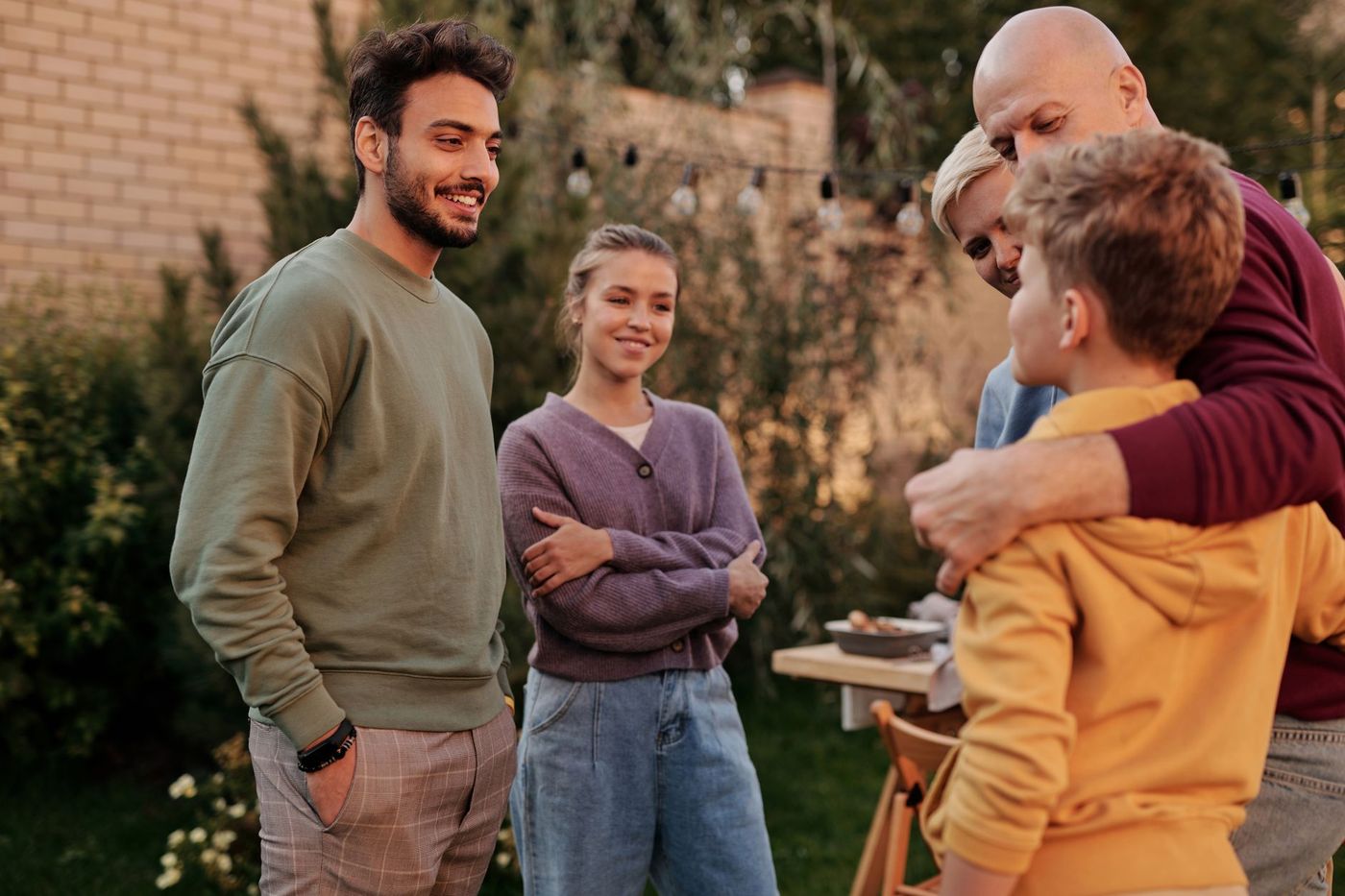 Grupo de cinco personas conversando y sonriendo al aire libre en un jardín, con una mesa y luces colgantes al fondo.