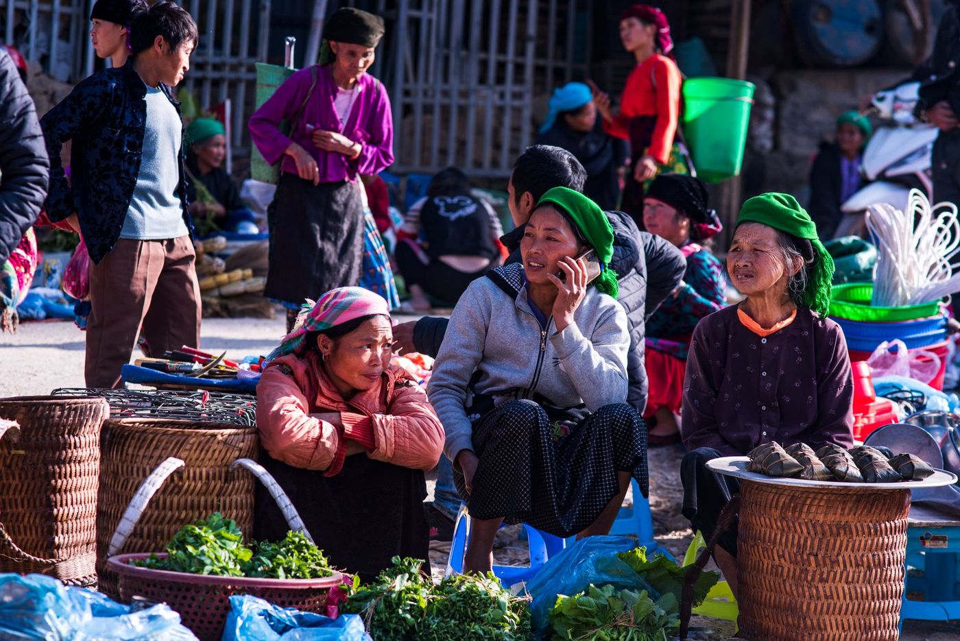Tres mujeres sentadas en un mercado al aire libre entre cestas de bambú, verduras frescas y alimentos, con otros vendedores alrededor.