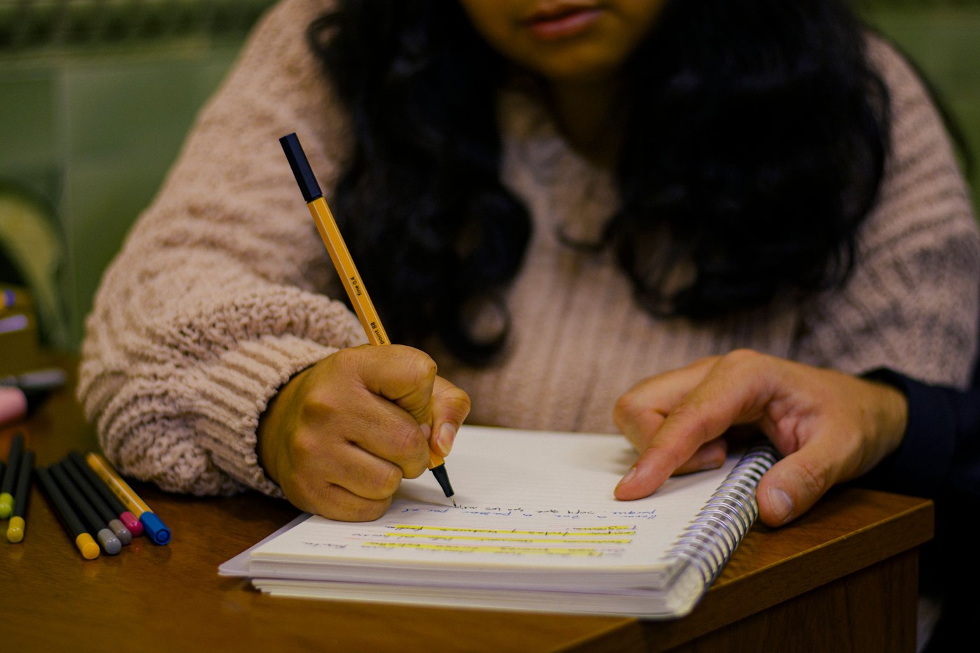 Primer plano de un estudiante escribiendo en un diario con bolígrafos de colores sobre un escritorio de madera.