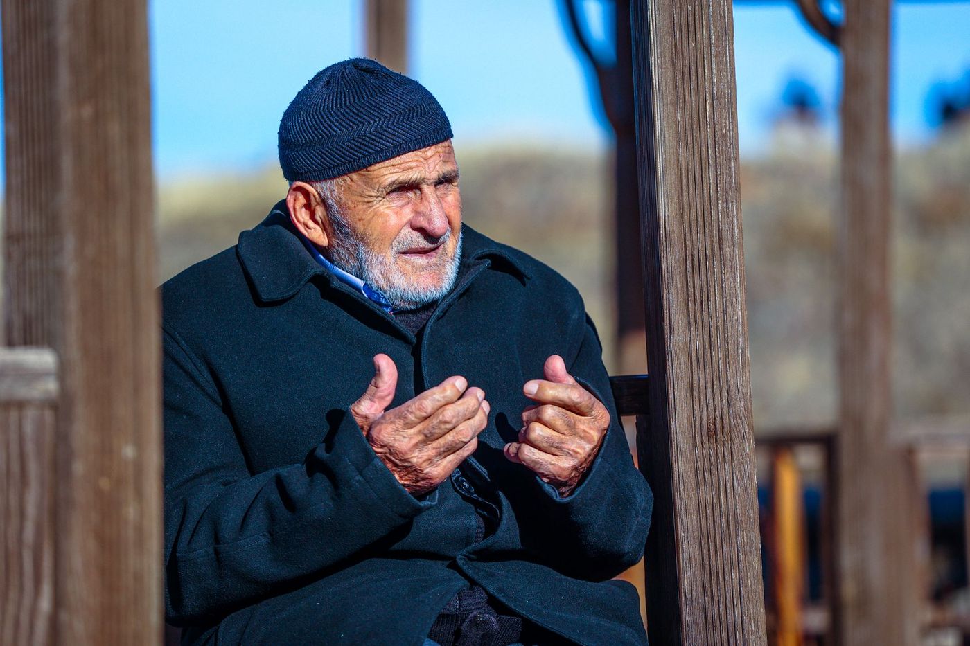 Un anciano de barba gris con gorro azul gesticula con las manos mientras conversa, sentado entre postes de madera.