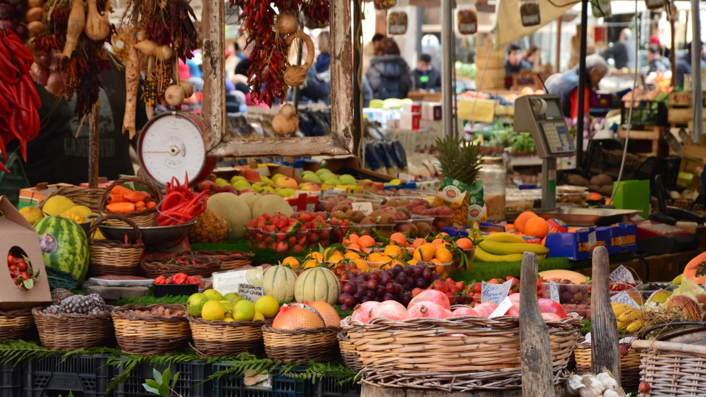 Escena colorida de mercado al aire libre mostrando frutas y verduras frescas en varios puestos.