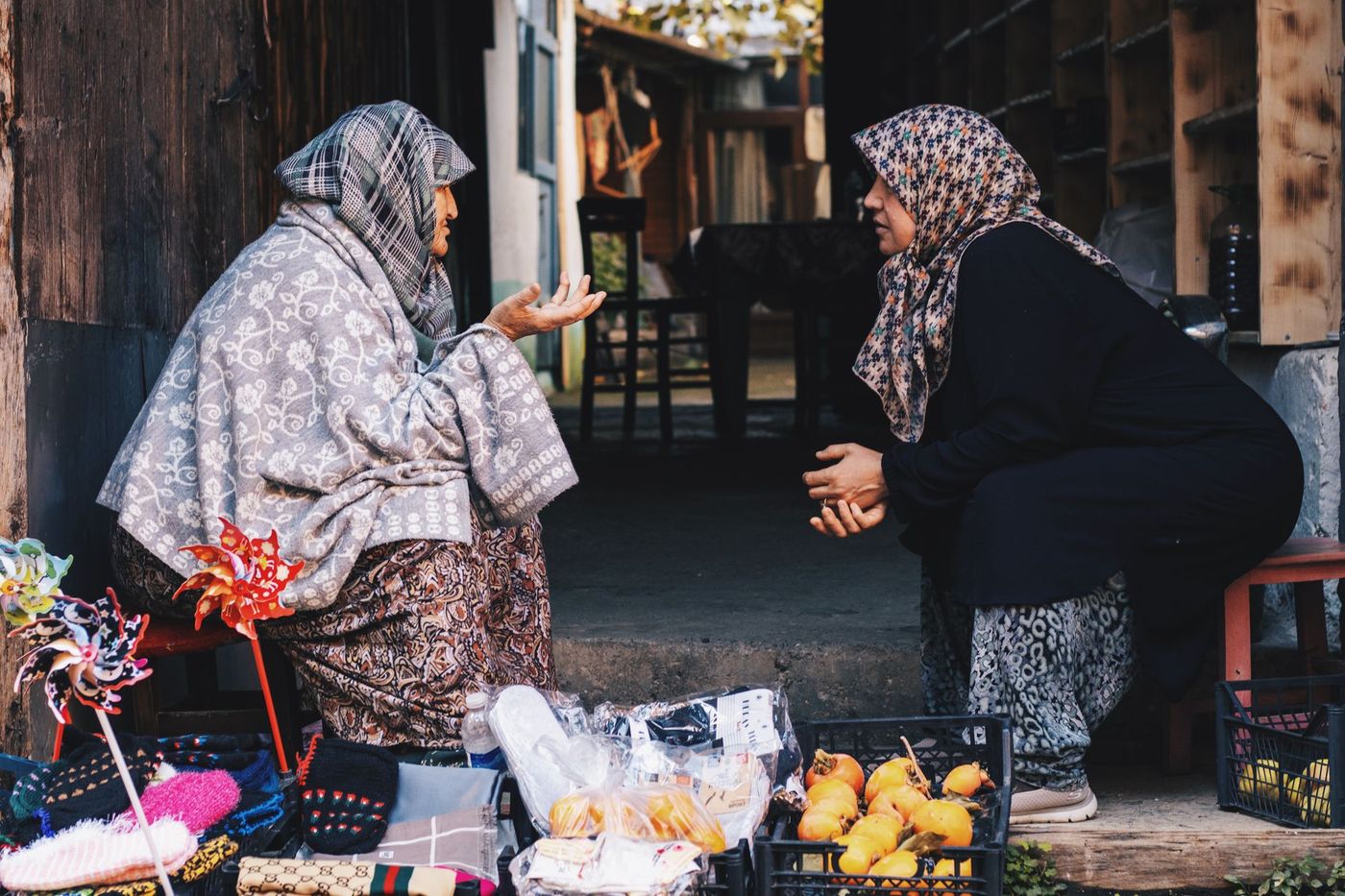 Dos mujeres conversando junto a un puesto callejero con artesanías, guantes de lana y cajas de fruta, en un entorno de madera.