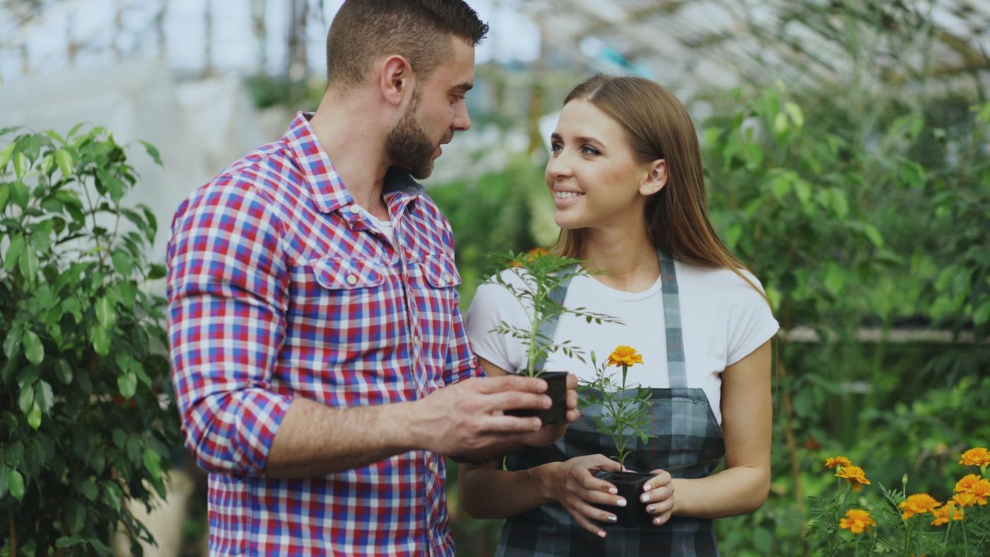 Pareja joven en un invernadero: él, con camisa de cuadros, sostiene una maceta con una planta, y ella, con delantal, sostiene otra maceta con una flor naranja mientras se miran sonrientes.