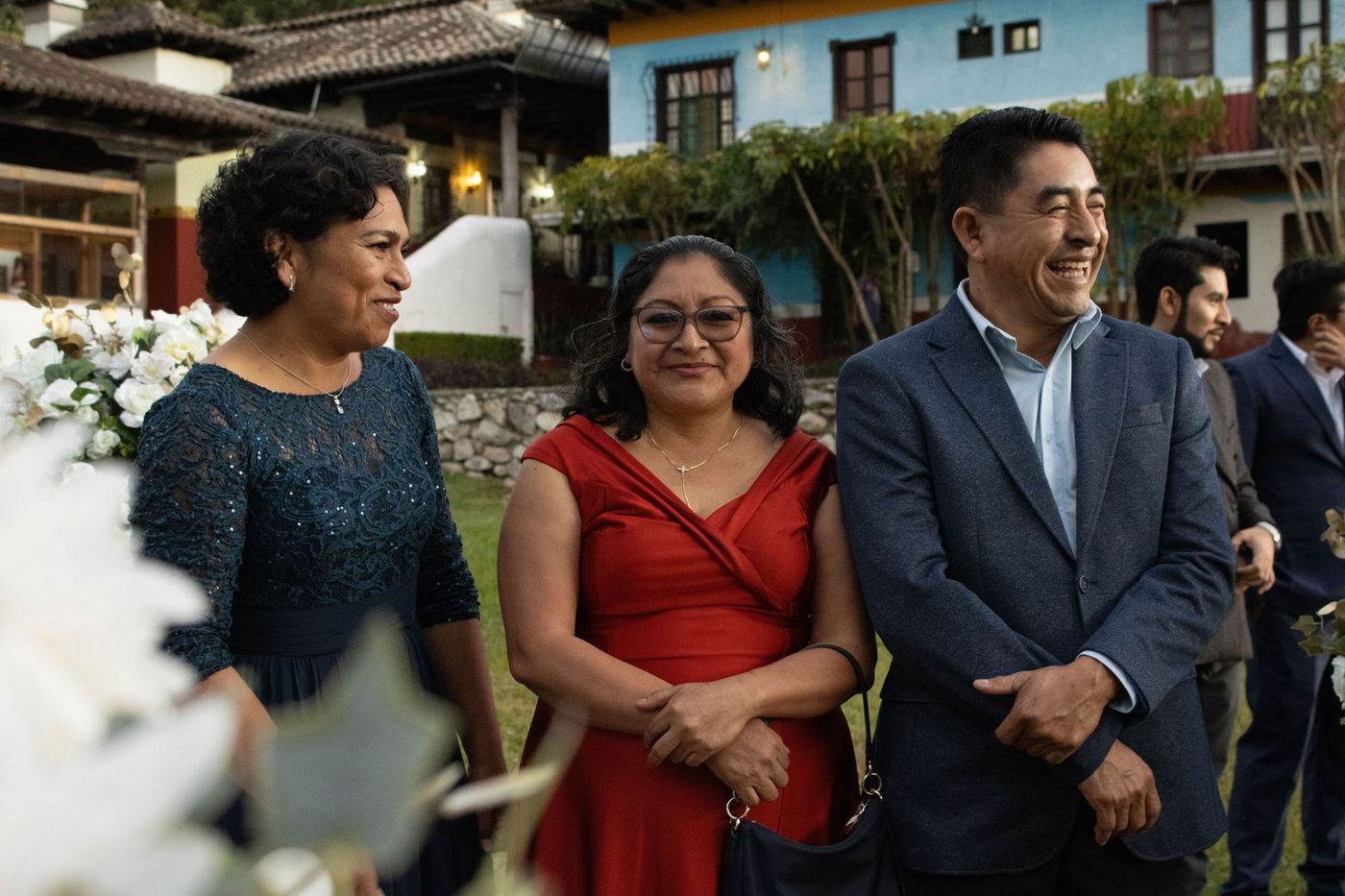Tres adultos elegantemente vestidos sonríen en un evento al aire libre, con flores blancas desenfocadas en primer plano y edificios, árboles e invitados al fondo.