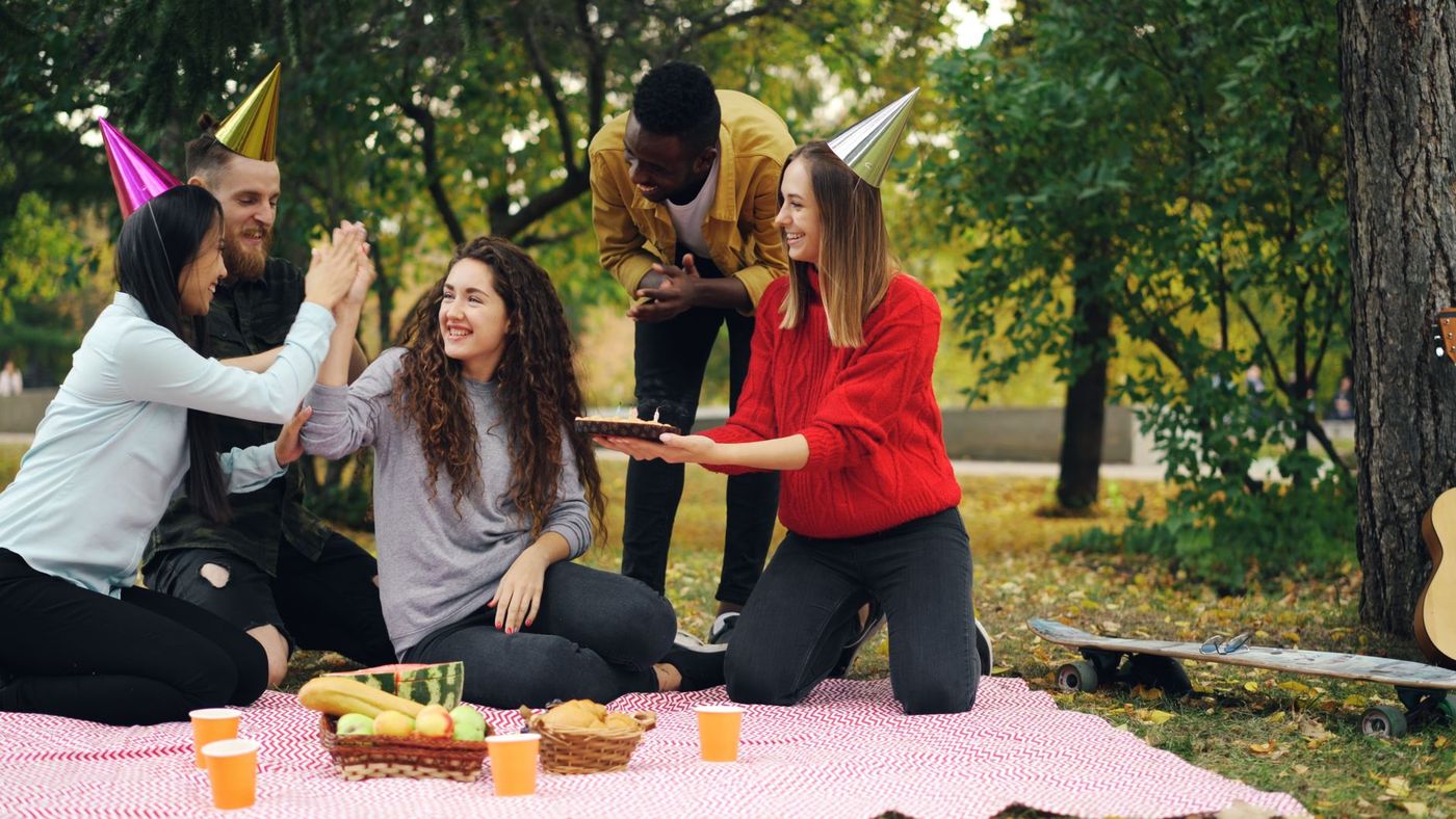 Cinco jóvenes celebran un cumpleaños en un picnic en el parque con gorros de fiesta, pastel con velas, fruta, cestas y una manta a cuadros.