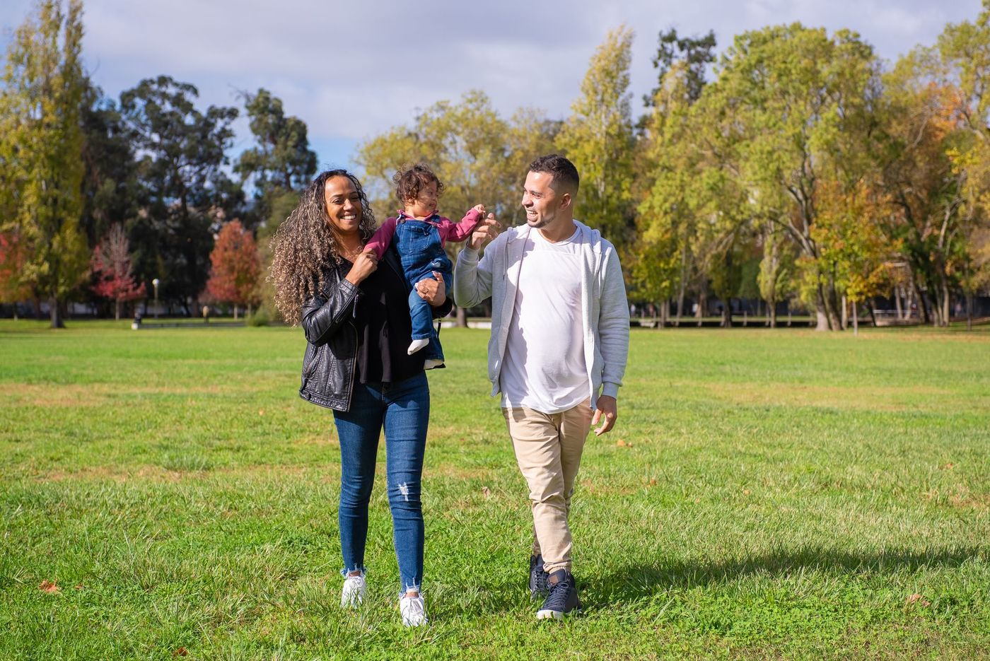 Familia de cuatro caminando sobre el césped de un parque soleado, con la mujer llevando una nevera portátil verde y los niños de la mano.