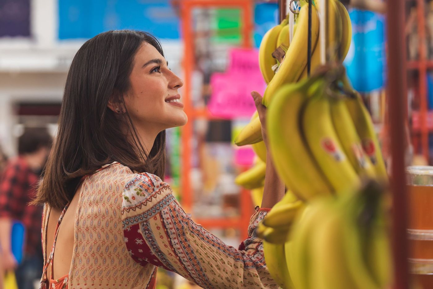 Mujer joven sonriente de perfil tocando y mirando racimos de plátanos colgantes en un mercado colorido con fondo desenfocado.