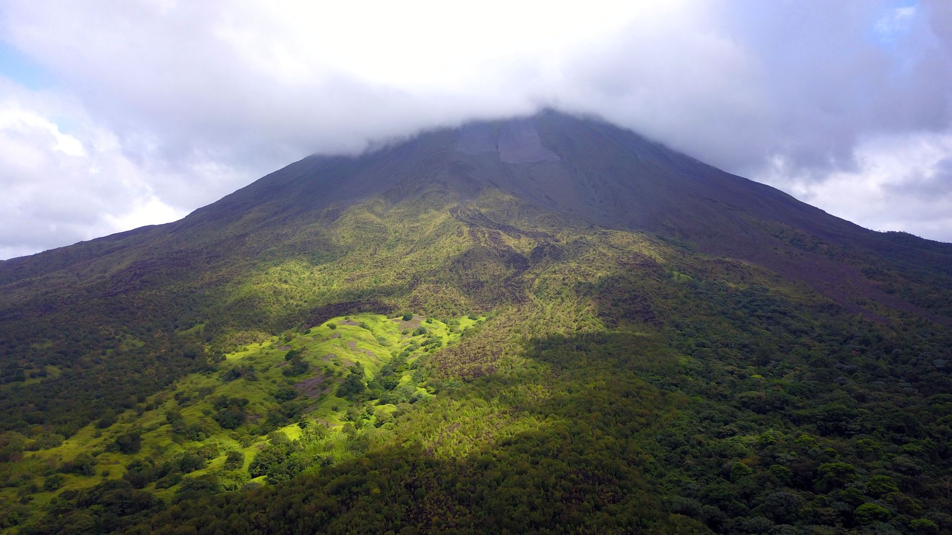 Volcán cónico con la cima cubierta por nubes y laderas verdes que descienden hacia un bosque denso, bajo un cielo parcialmente nublado.