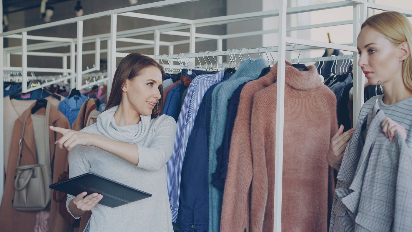 Dos mujeres comprando ropa, una sosteniendo una tableta, discutiendo opciones en una tienda.