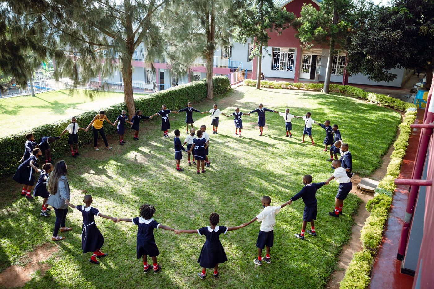 Vista elevada de un grupo de niños y niñas con uniforme escolar que, tomados de la mano y acompañados por dos adultas, forman un círculo en un patio de césped rodeado de árboles.