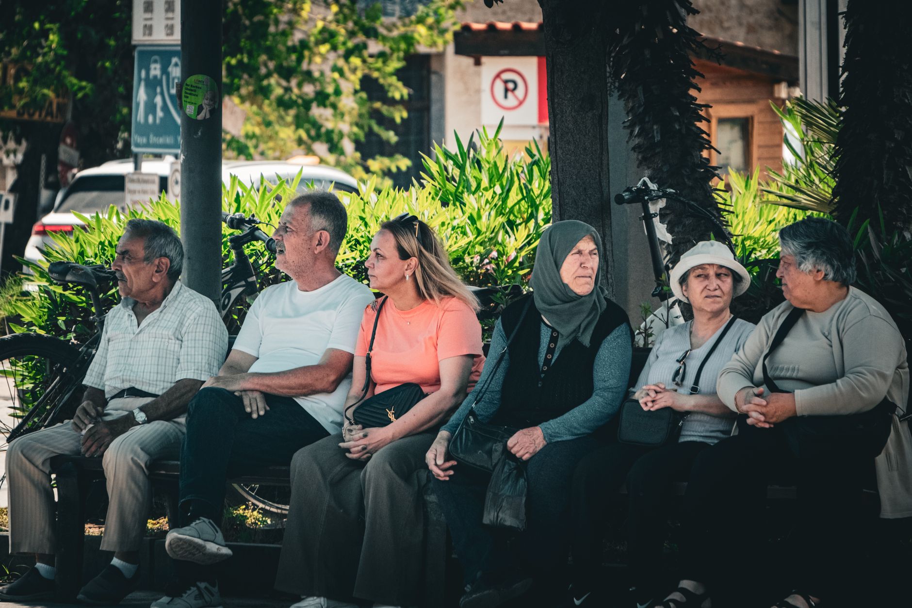 Seis personas sentadas en un banco público al aire libre, con bicicletas, plantas y señales de tránsito alrededor, en una escena urbana cotidiana.