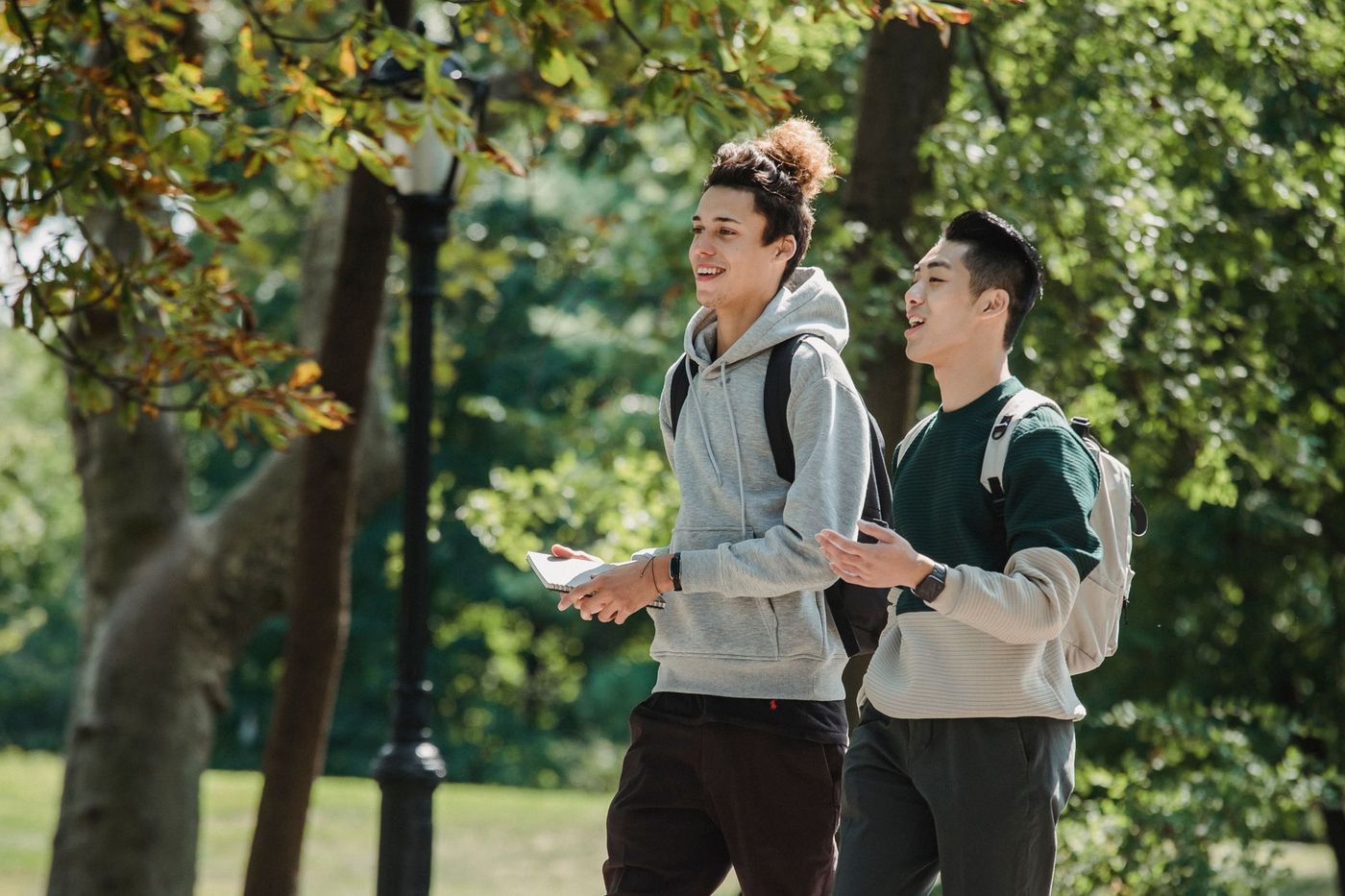 Dos jóvenes estudiantes con mochilas caminan y conversan sonrientes por un sendero arbolado y soleado.