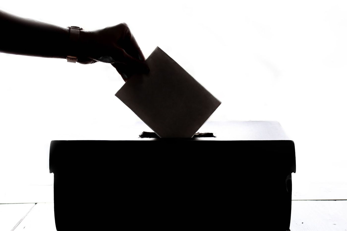 Silueta de una mano depositando una papeleta en la ranura de una urna electoral, contra un fondo blanco iluminado.