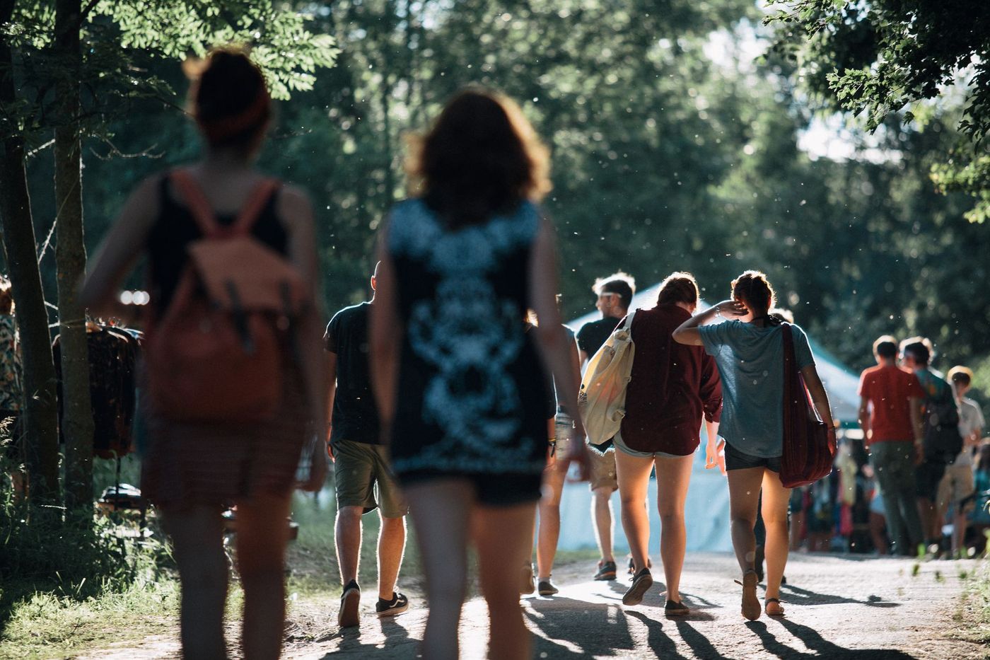 Grupo de jóvenes caminando por un sendero de tierra entre árboles hacia una carpa blanca, con luz solar filtrada y polvo en el aire.