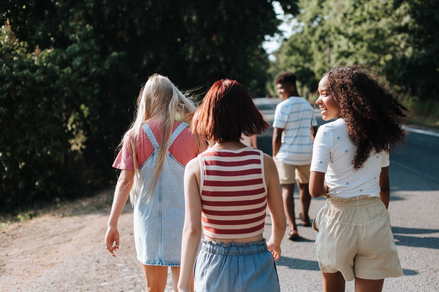Tres chicas y un chico caminan de espaldas por un camino arbolado y soleado, con una de las chicas volteando sonriente hacia atrás.