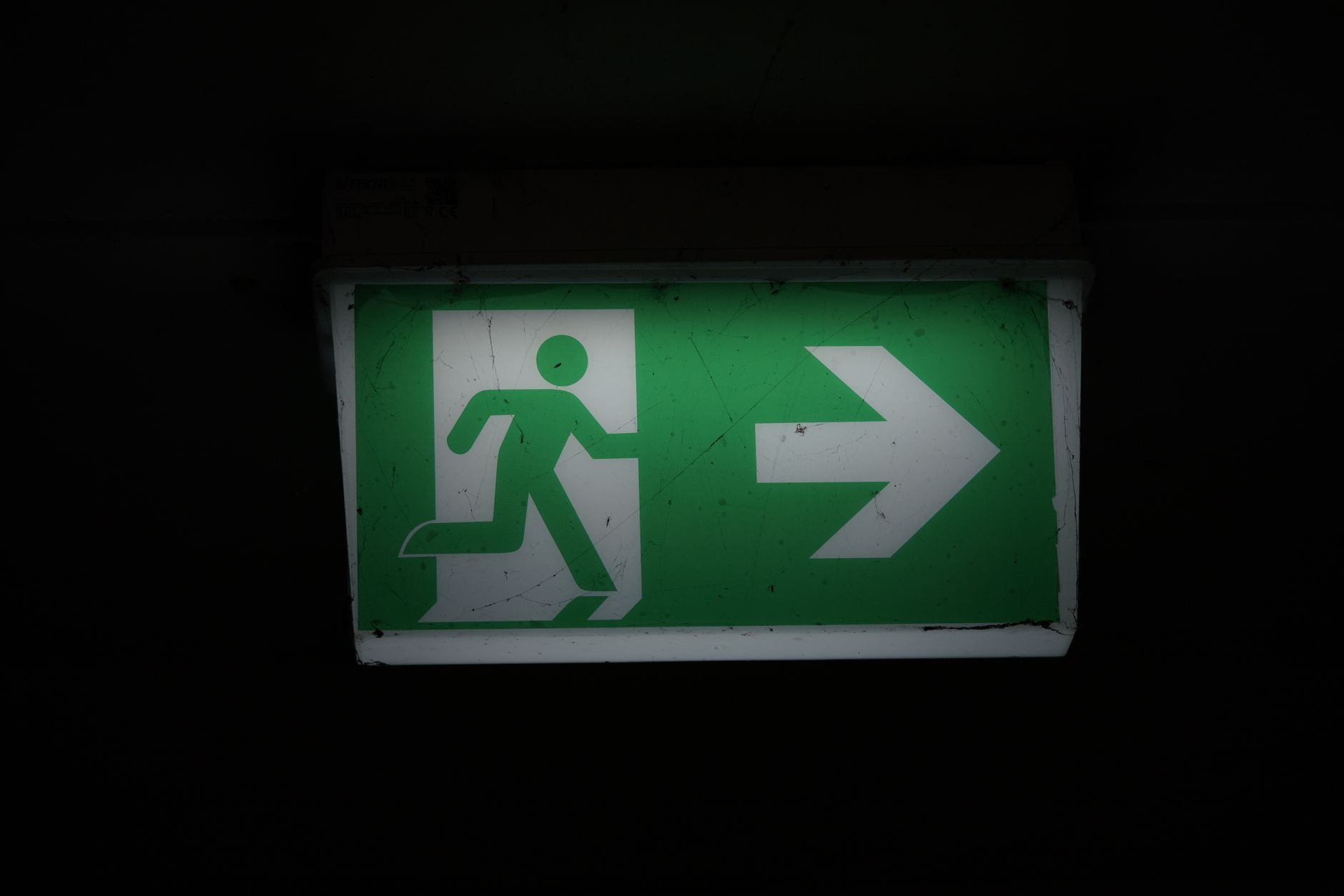 Señal de salida de emergencia iluminada en verde, con el pictograma blanco de una persona corriendo y una flecha que apunta a la derecha, sobre un fondo oscuro.