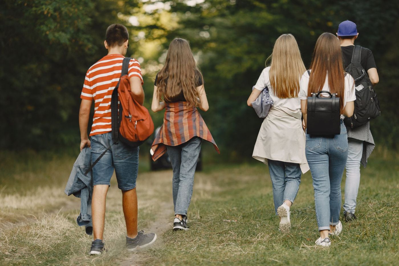 Cinco jóvenes con mochilas y ropa casual caminan de espaldas por un sendero rodeado de árboles en un día soleado.