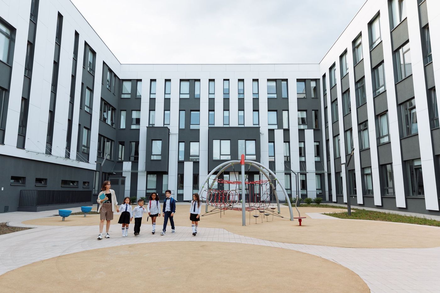 Patio escolar moderno con una maestra y cinco niños uniformados caminando junto a un juego de trepa metálico con redes rojas, rodeado de edificios blancos y grises.