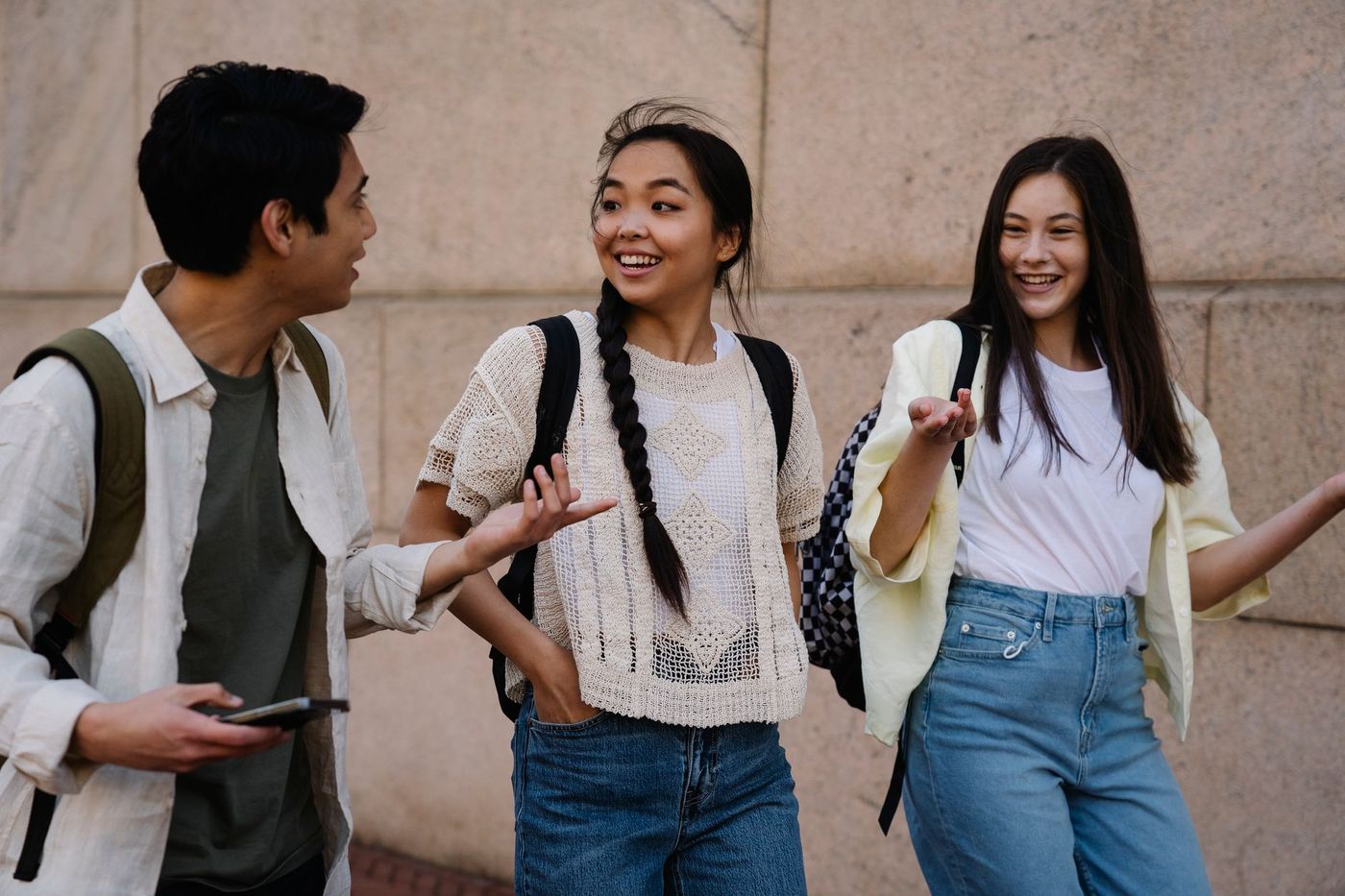 Tres jóvenes con mochilas conversan y gesticulan sonrientes junto a una pared de piedra beige, con ropa casual y jeans.