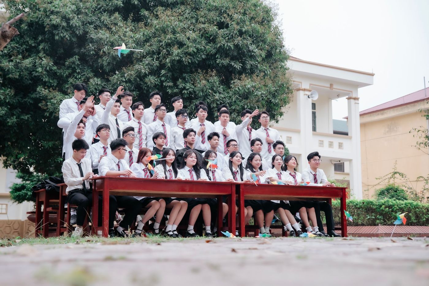 Estudiantes adolescentes con uniforme de camisa blanca y corbata roja posan al aire libre sobre mesas de madera roja, con molinetes de papel y un gran árbol de fondo.