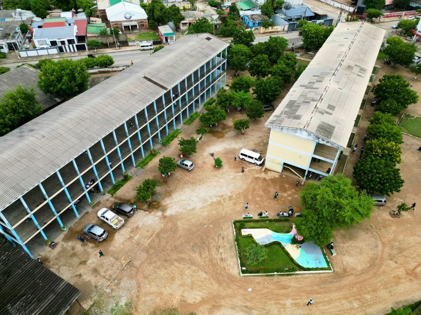 Vista aérea de un patio escolar de tierra con dos largos edificios de aulas, vehículos y árboles, rodeado por un barrio urbano.