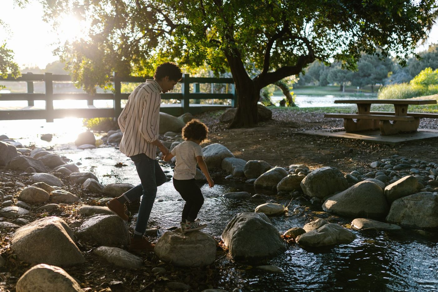 Un adulto y un niño caminan de la mano sobre las rocas de un arroyo dentro de un parque arbolado, con una mesa de picnic y una valla de madera al fondo.