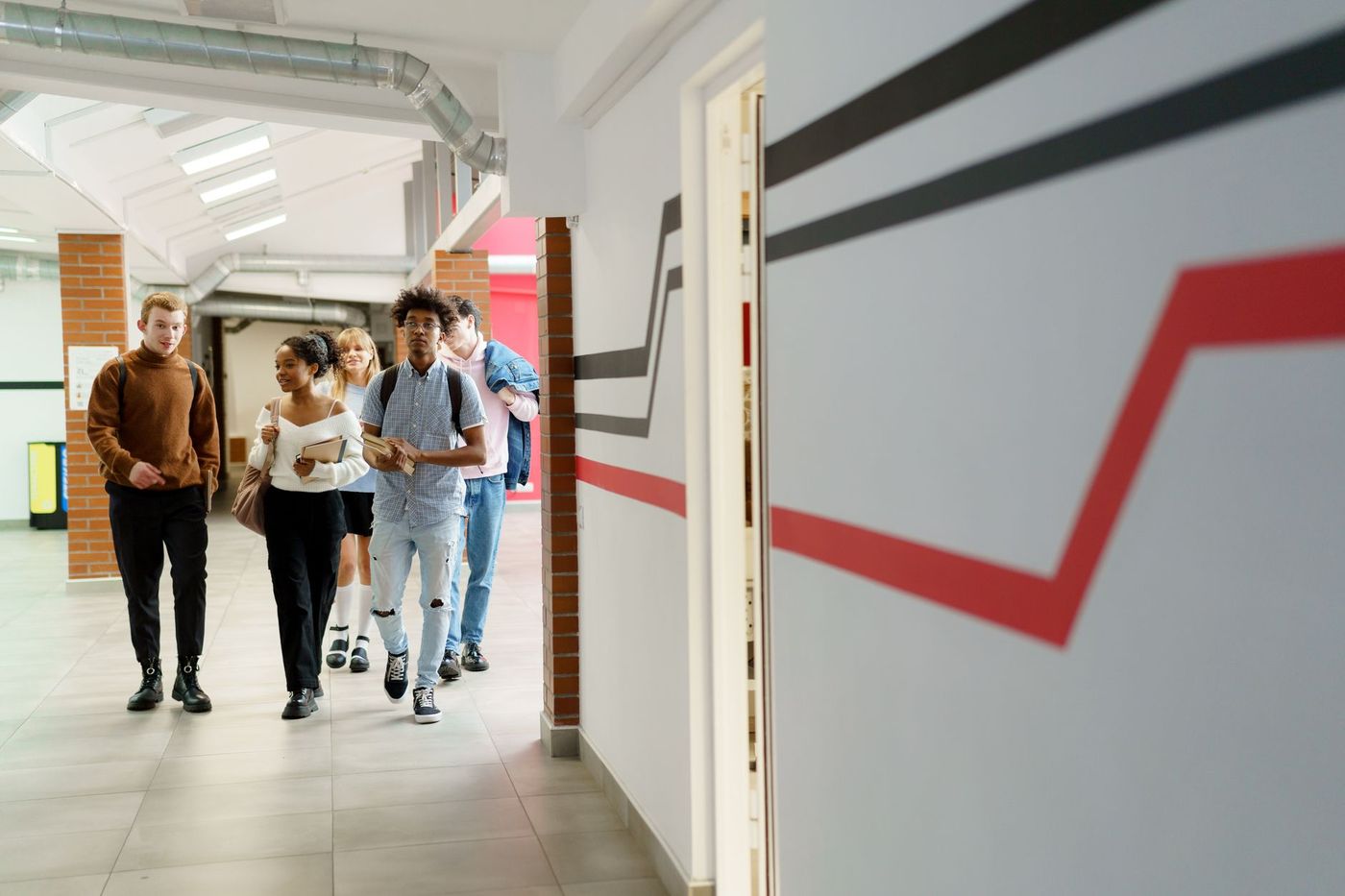 Cinco estudiantes jóvenes caminan por un pasillo escolar moderno con columnas de ladrillo y techos con conductos metálicos.