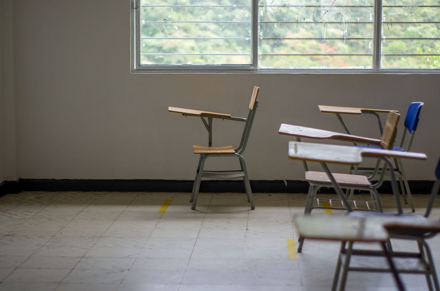 Aula escolar vacía con pupitres individuales separados, marcas amarillas en el suelo y una ventana grande con vista a los árboles.