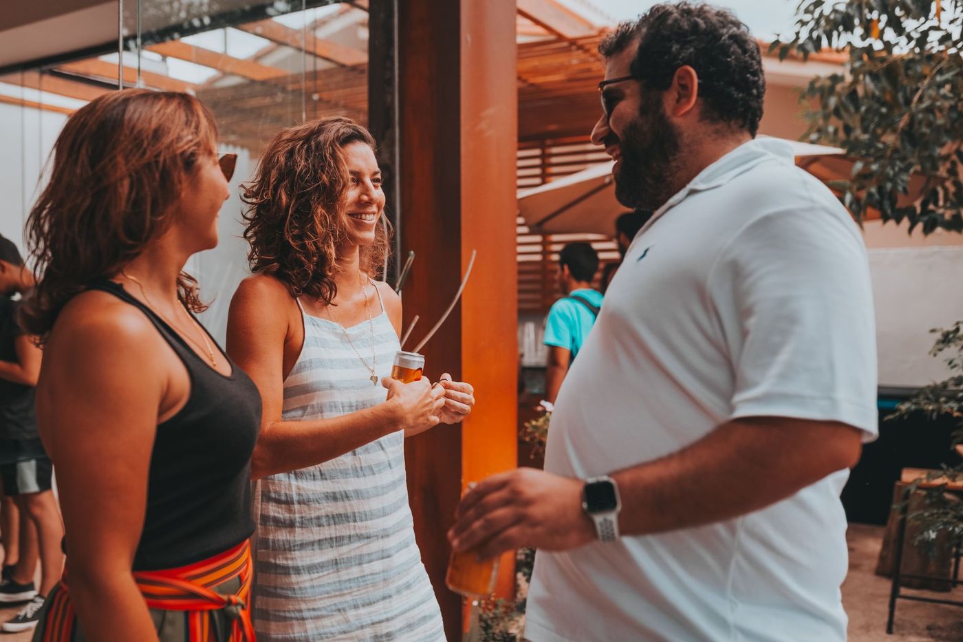 Tres adultos conversan y ríen con bebidas en la mano en una terraza con estructura de madera, cristal y plantas.