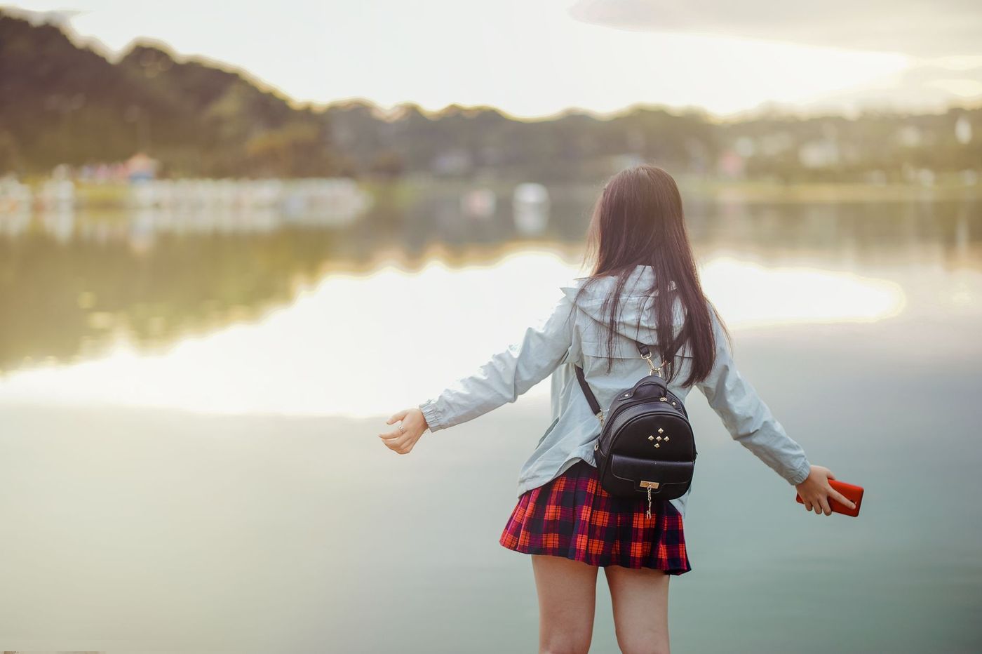 Mujer joven de espaldas, con chaqueta clara y falda escocesa, abre los brazos frente a un lago tranquilo al atardecer.