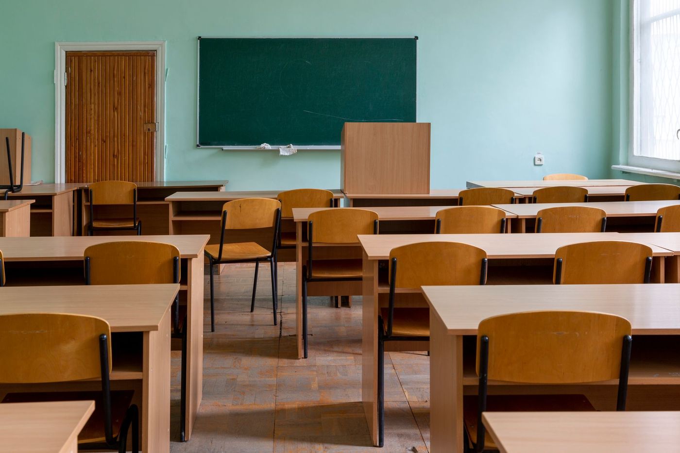 Aula escolar vacía con pizarra verde al fondo, filas de pupitres y sillas de madera clara, paredes verde menta y ventana con luz natural.