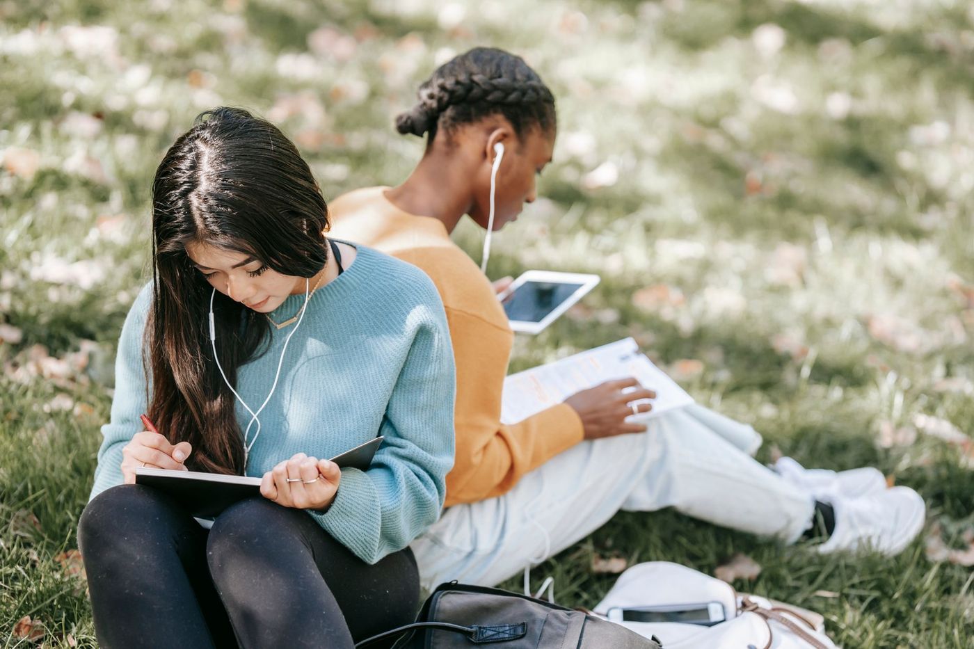 Dos jóvenes estudiantes sentadas en el césped, una escribiendo en un cuaderno y otra consultando una tableta, ambas con auriculares blancos.