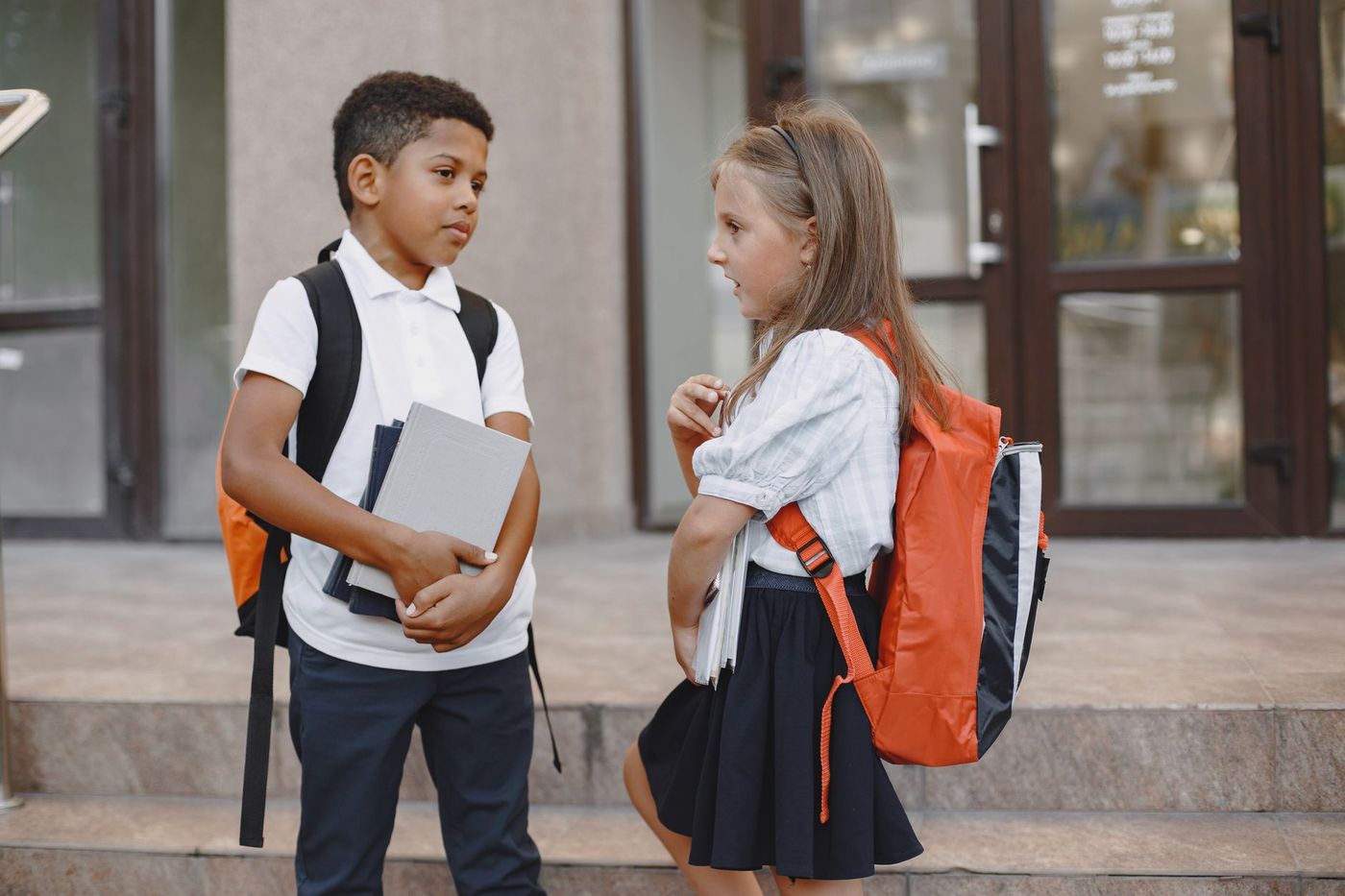 Dos niños escolares con mochilas y cuadernos conversan de pie en las escaleras de la entrada acristalada de un edificio.