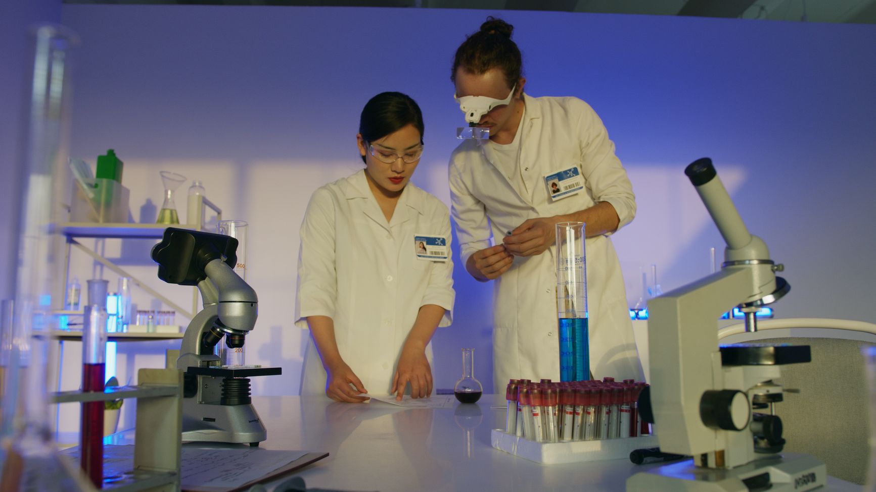 dos investigadoras con bata blanca trabajan en el laboratorio con microscopios, tubos y matraces