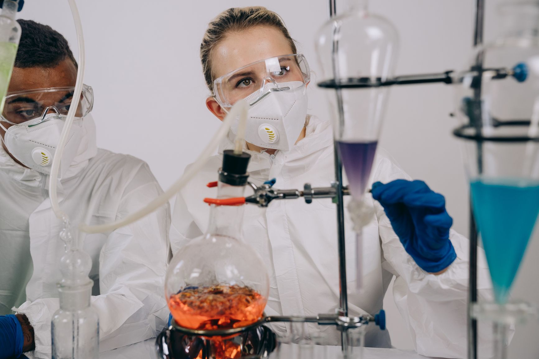 dos personas con trajes de protección, mascarilla, gafas y guantes manipulan material de vidrio con líquidos en el laboratorio