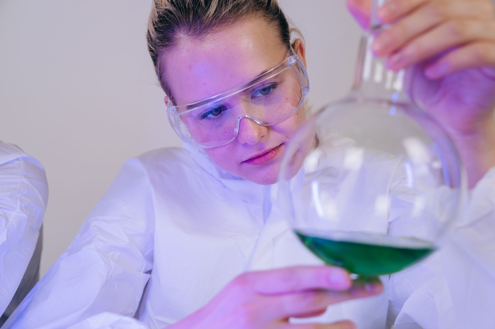 una joven con gafas de seguridad y bata blanca examina un matraz con disolución verde en el laboratorio