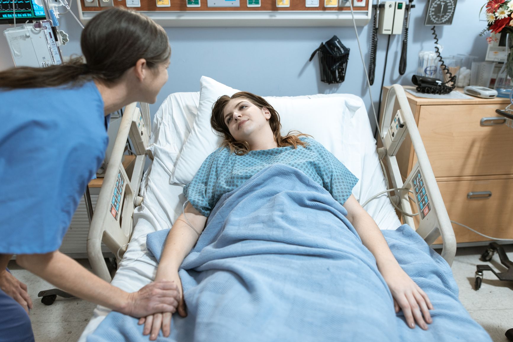 una profesional de la salud con scrubs azules sostiene la mano de una paciente con vía intravenosa en una habitación de hospital
