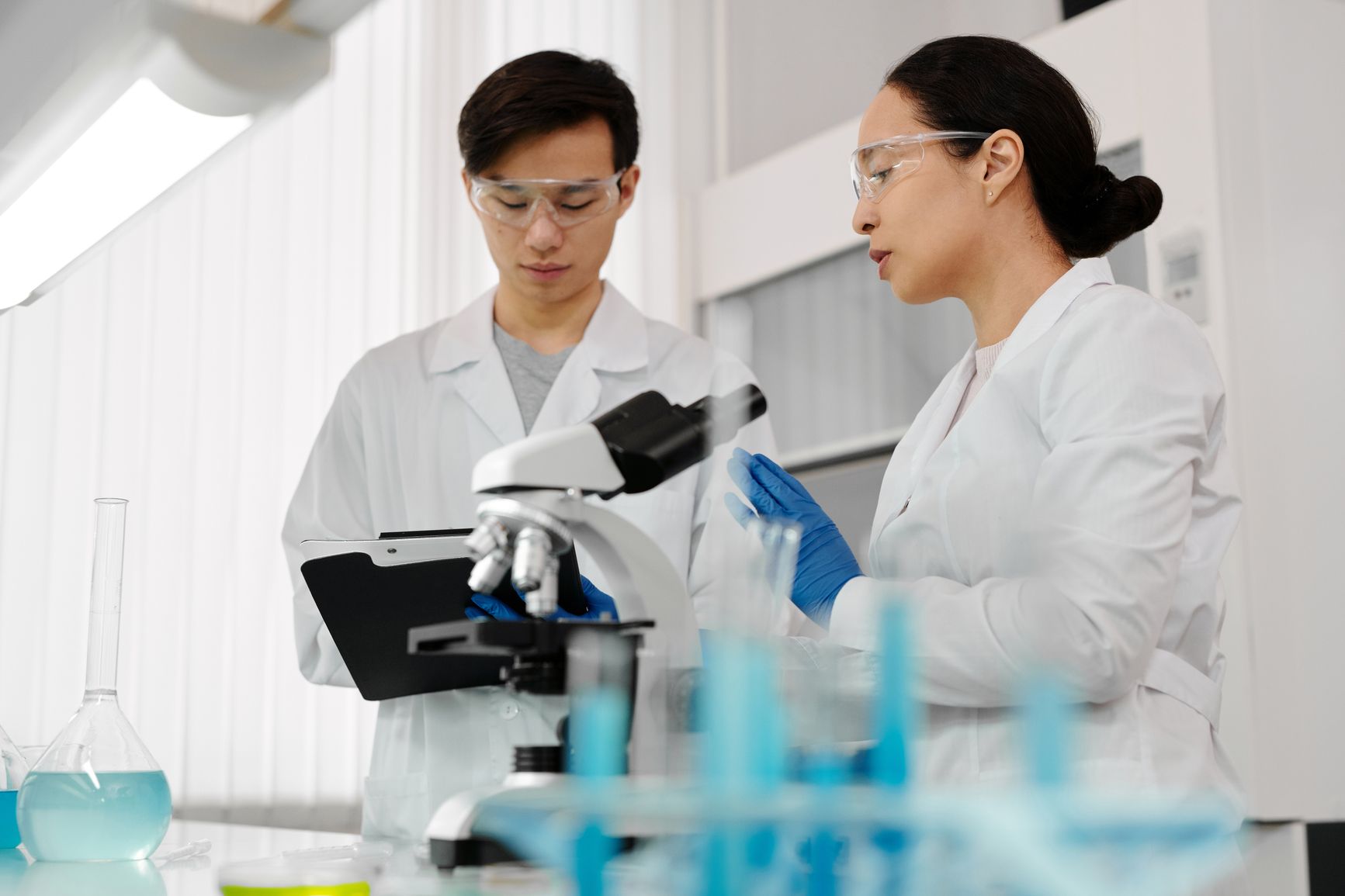 dos personas con bata y gafas de laboratorio examinan un microscopio y revisan anotaciones junto a tubos y un matraz