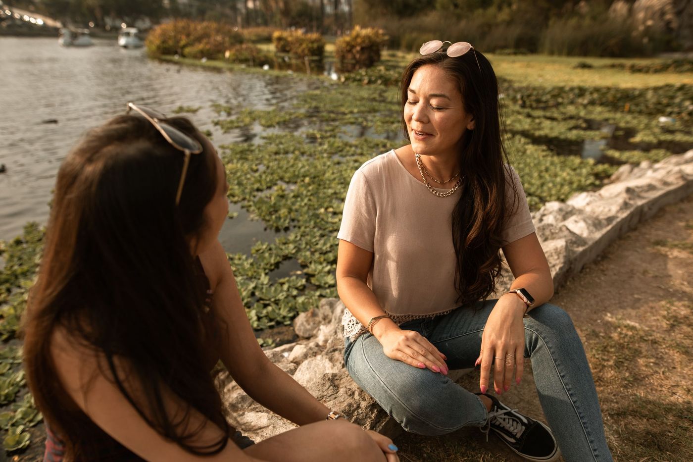 dos mujeres adultas conversan sentadas junto al agua, rodeadas de vegetación