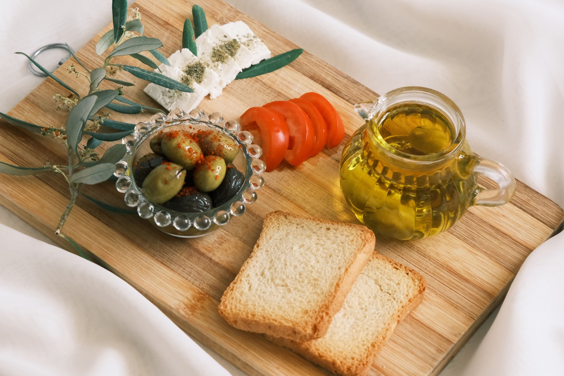 Tabla de madera con pan, aceitunas, tomate, queso y aceite de oliva sobre tela blanca.