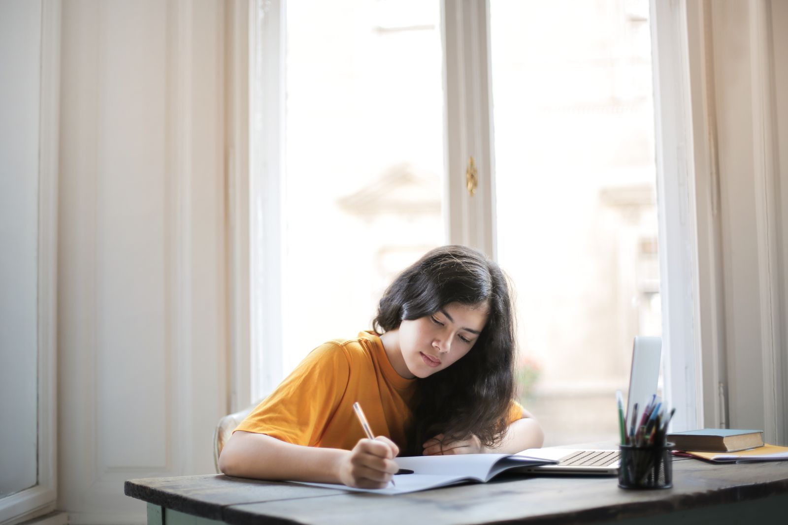 Estudiante escribiendo en un cuaderno abierto con un bolígrafo, junto a una ventana con luz natural