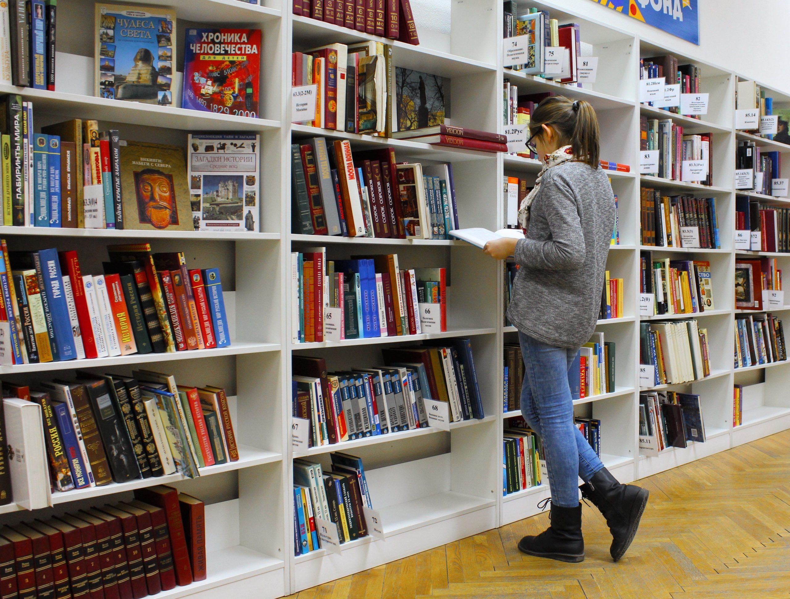 Una mujer joven con gafas y suéter gris lee un libro abierto de pie en un pasillo entre estanterías blancas de una biblioteca.