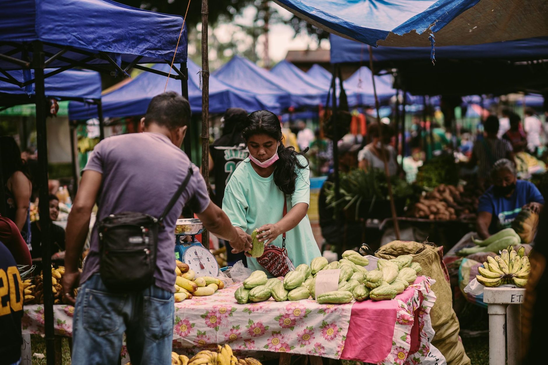 Puesto de frutas y verduras en un mercado al aire libre: una vendedora tras la mesa, con la balanza y los chayotes apilados, y un cliente de espaldas frente al puesto.
