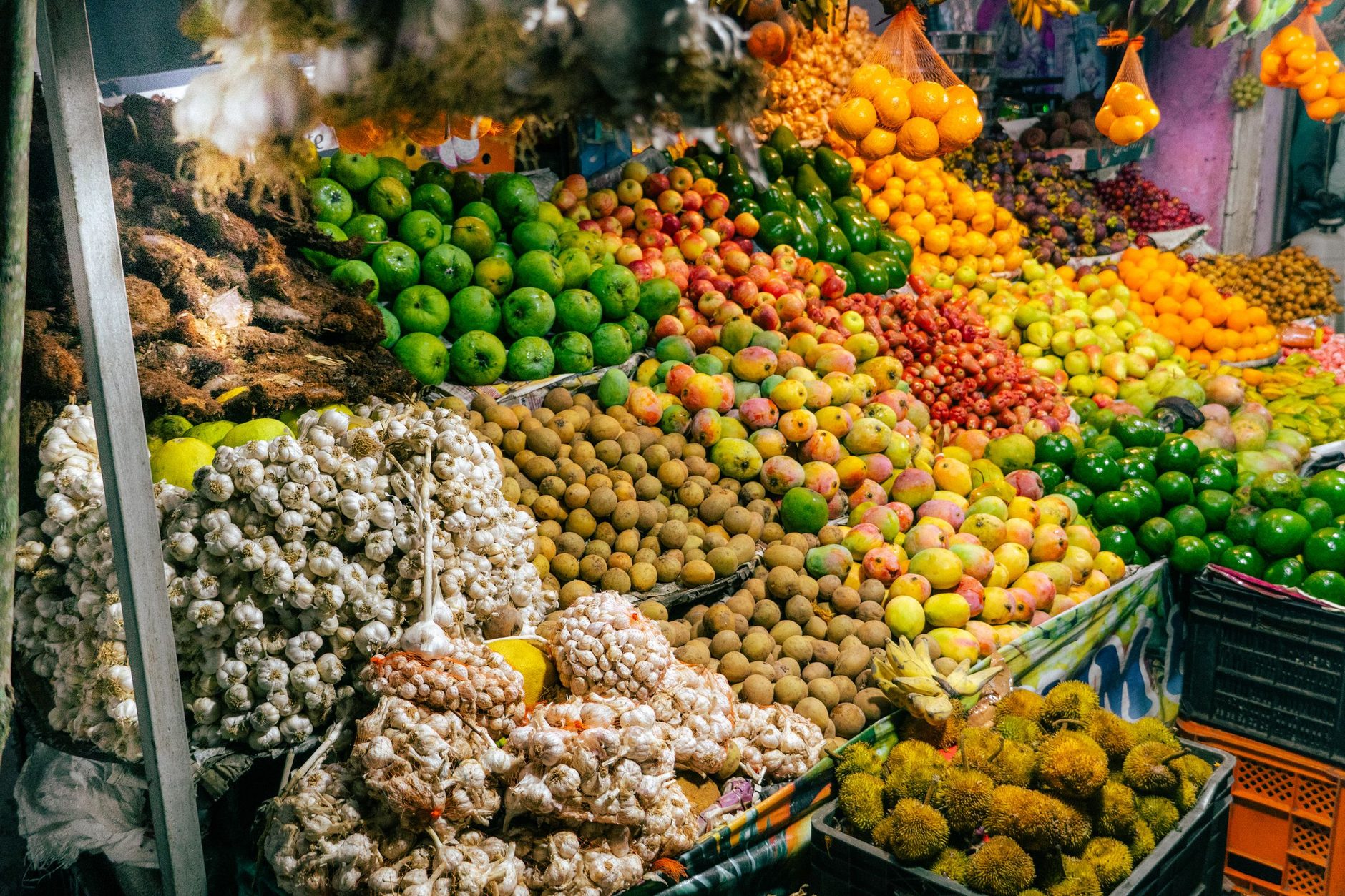 Puesto de frutas y verduras al aire libre con pilas de ajos, tubérculos y frutas en cajas, y racimos de plátanos colgando bajo el techo.