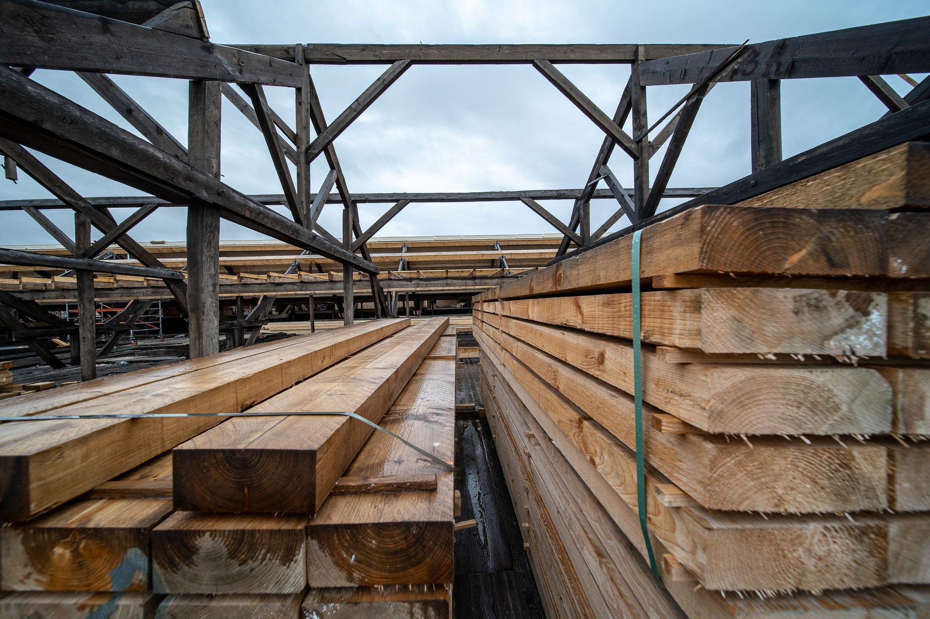 Tablones de madera apilados en un patio al aire libre, con una estructura de vigas de madera al fondo.