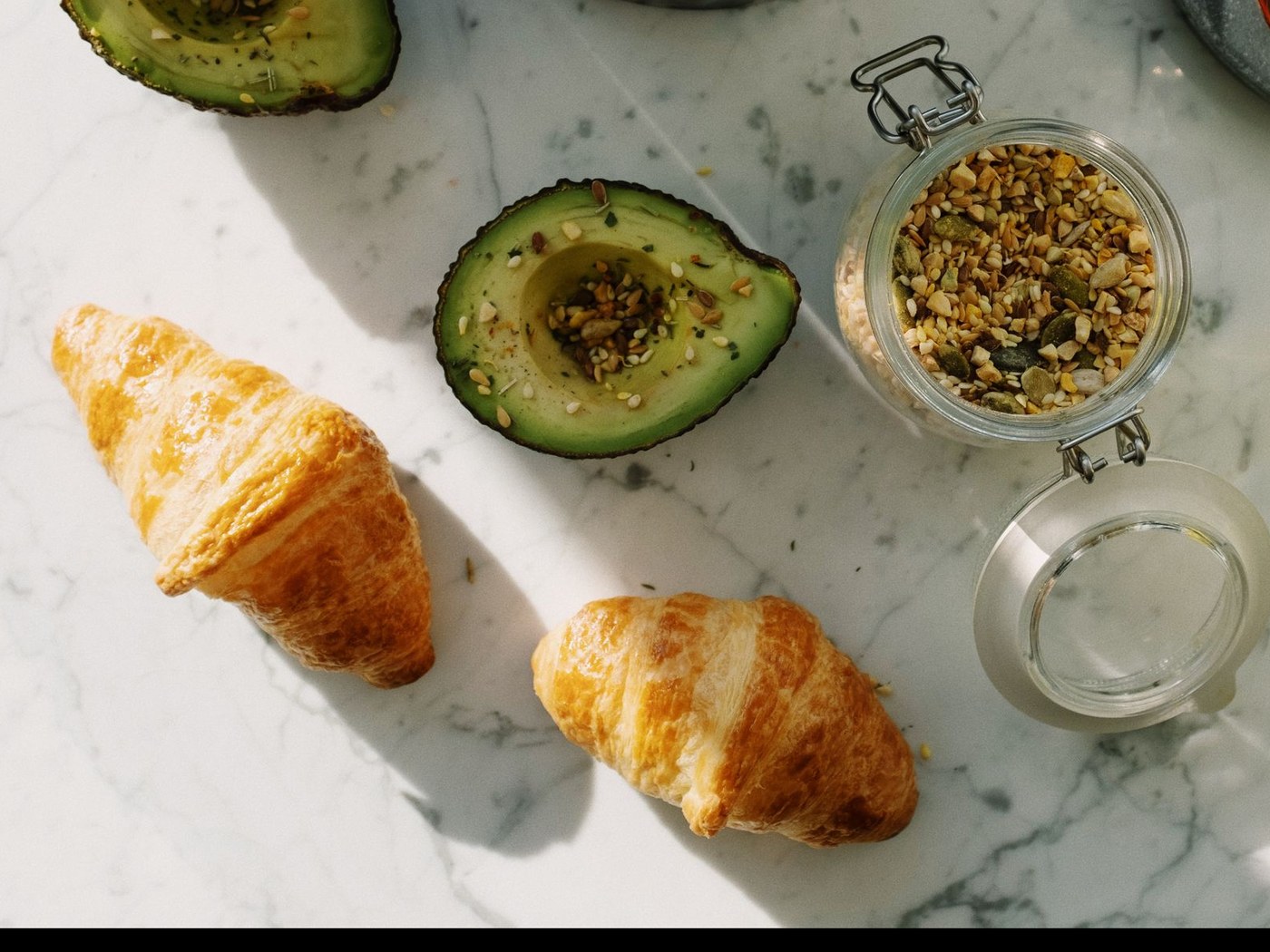 Dos croissants dorados, dos medios aguacates cubiertos de semillas y un frasco de semillas sobre una mesa de mármol blanco.