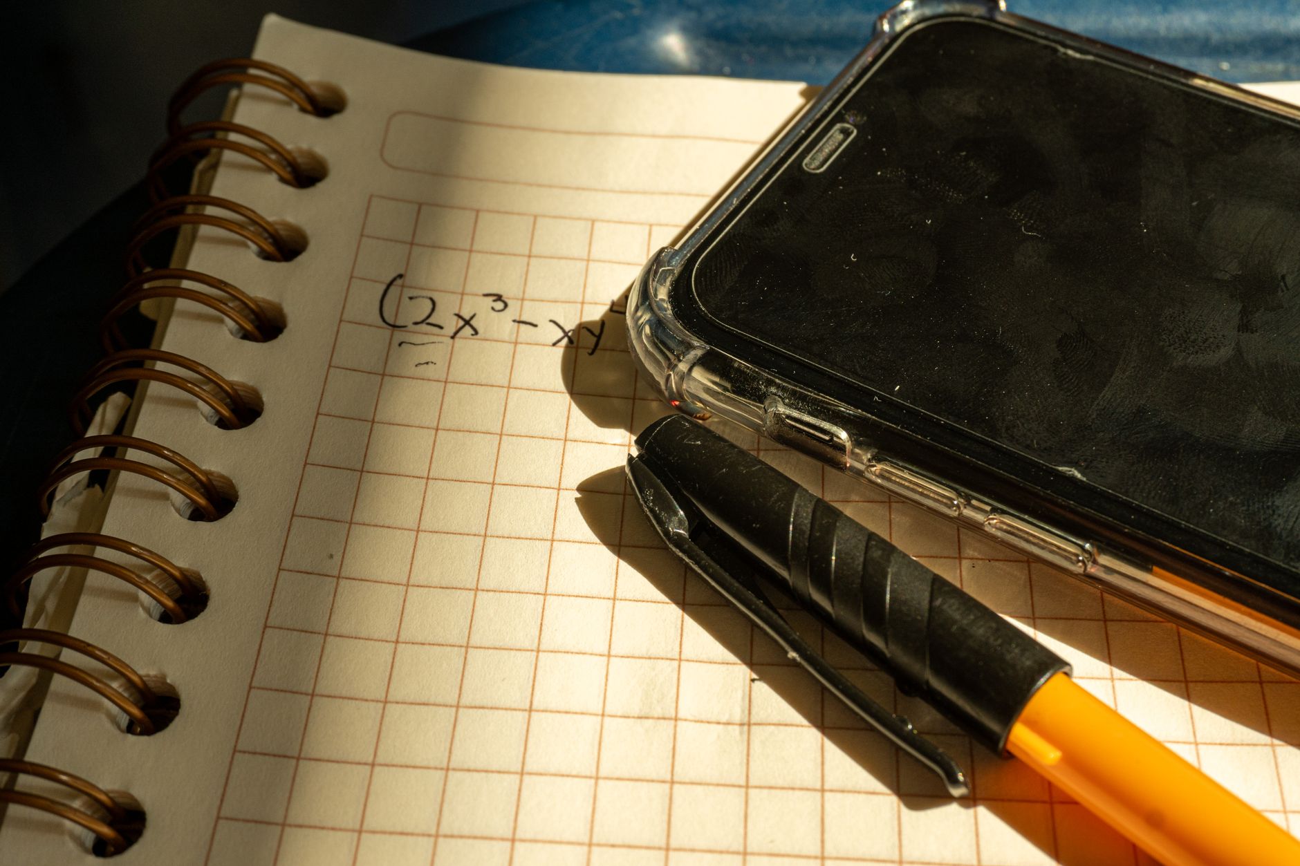 Cuaderno cuadriculado con la expresión algebraica dos equis al cubo menos equis escrita a mano, junto a un bolígrafo y un teléfono sobre una superficie oscura