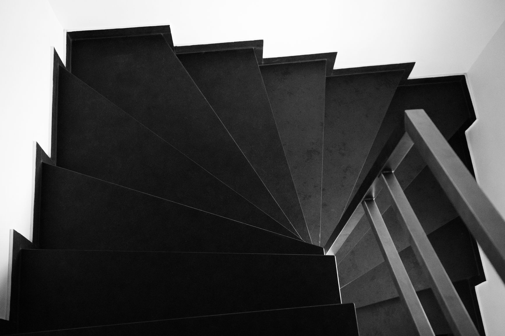 Escalera de caracol fotografiada en blanco y negro desde abajo, con los peldaños en forma de cuña que giran alrededor de un eje vertical y una barandilla gris a la derecha