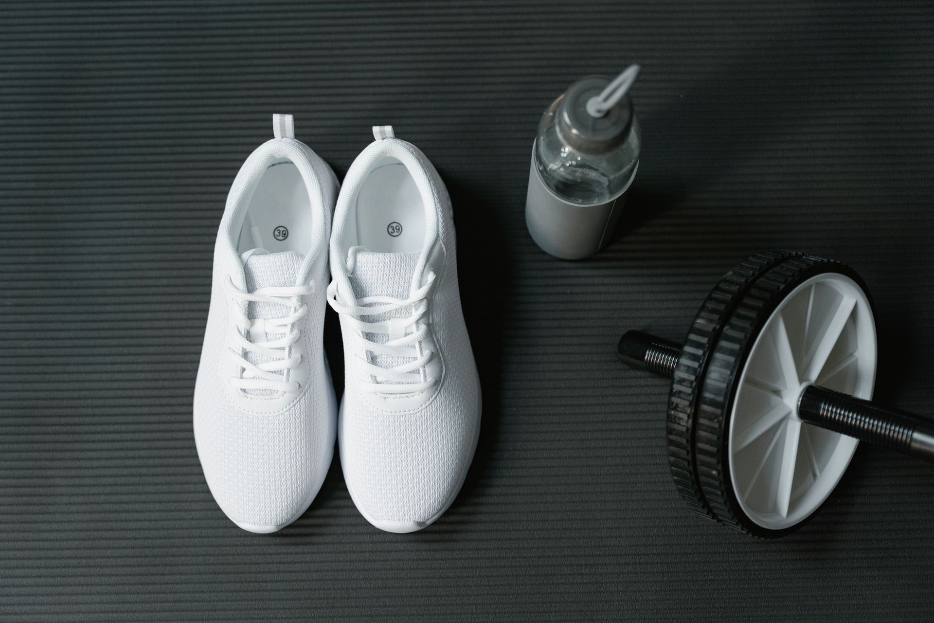 Zapatillas deportivas blancas y una botella de agua sobre una esterilla oscura, junto a una rueda de abdominales