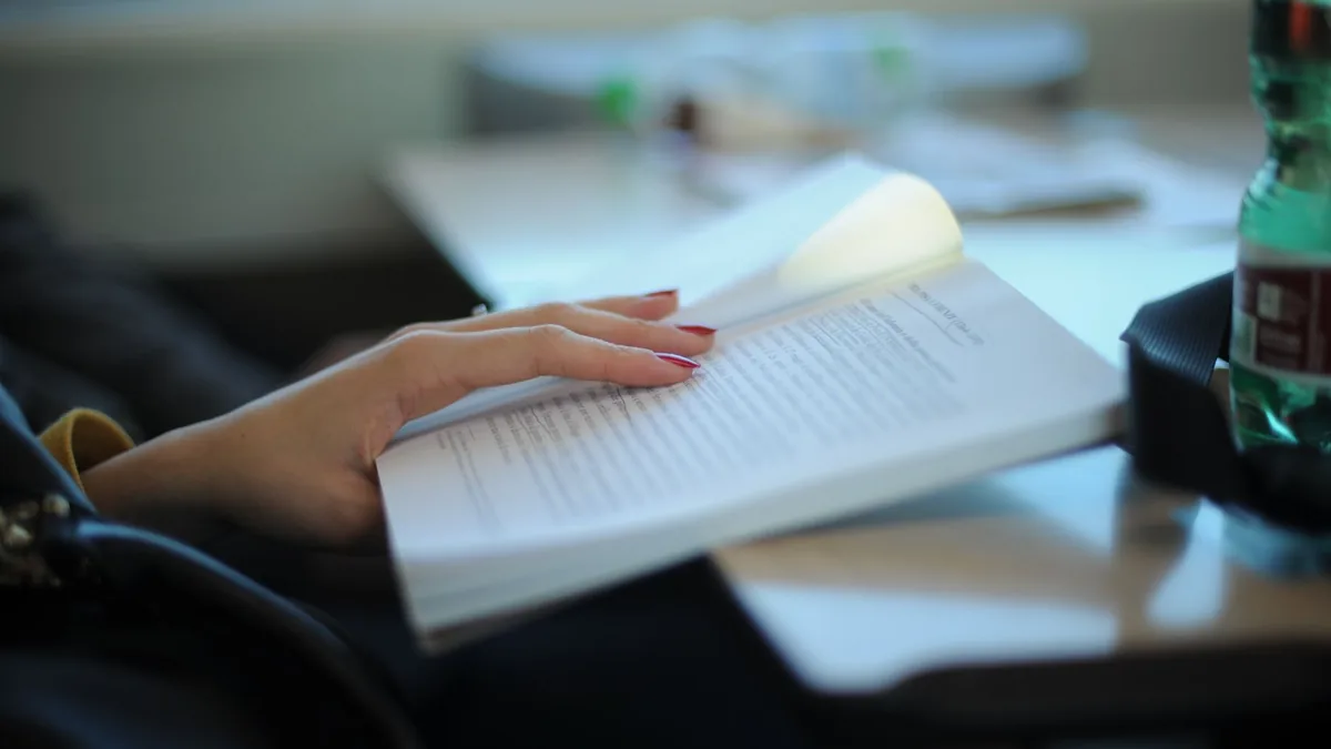 Una mujer con uñas arregladas está leyendo un libro en un escritorio, enfatizando el estudio y la relajación.